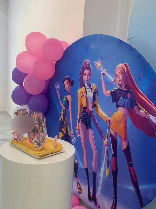 Bolo de aniversário numa coluna branca, ao lado de um painel com personagens de desenhos animados e balões cor-de-rosa e roxos.