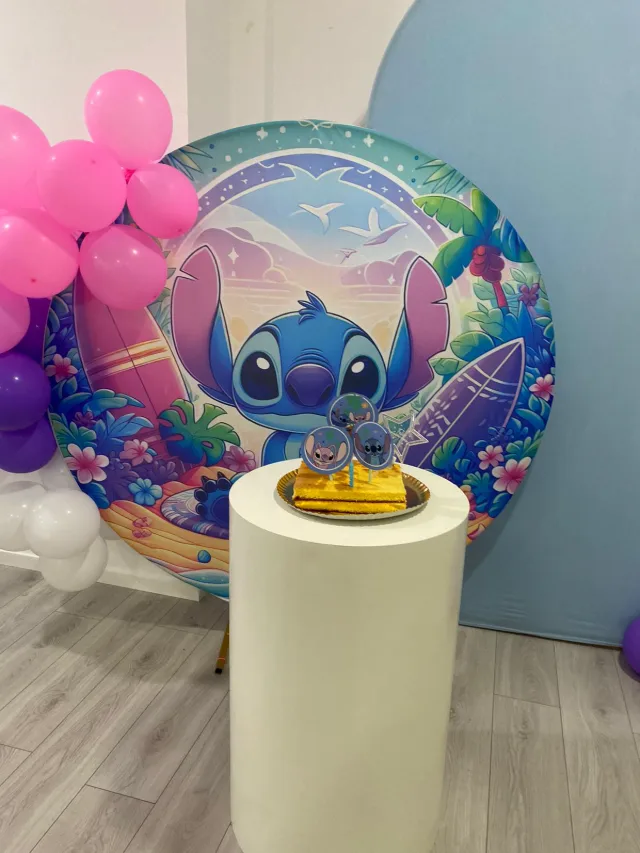 Decoração de festa com painel redondo do Stitch, balões cor-de-rosa e o bolo numa coluna branca.