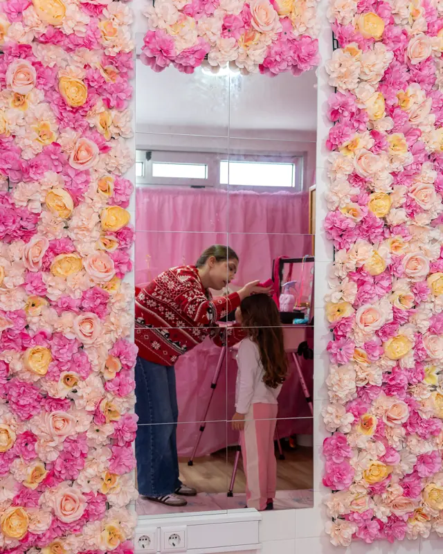 Salão de spa cor-de-rosa com parede de flores e uma menina a ser maquilhada.